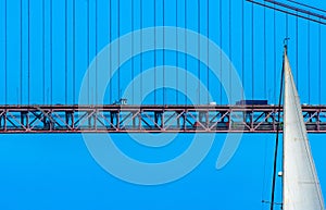 Top of the white sail of a sailboat sailing in front of the red steel 25 de Abril suspension bridge with road traffic and a train