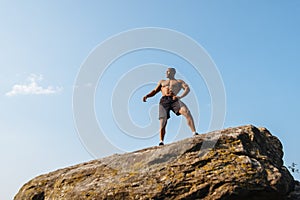 Topless portrait of strong black african american man bodybuilder posing on the rock. Blue cloudy sky background