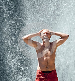 Polonahá upřímně smějící se muž stojící pod vodopádem horské řeky a užívající si stříkající přírodní sílu. Středního věku