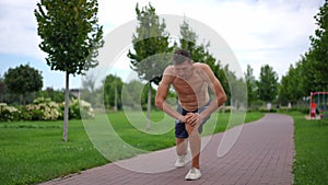 Topless sportsman in shorts stretching leg muscles in slow motion on park alley. Wide shot portrait of fit athletic