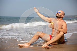 Topless vacationer in sunglasses with drink in hand lifting arm up and enjoying sun with closed eyes on wet seashore.