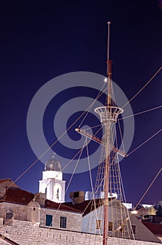Topmast and cables of an old ship from the time of the discoveries against a clear bell tower at night.
