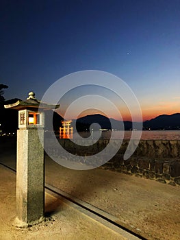 Tori gate in the middle of the ocean at the background, during sunset, with a post lamp shining on the first plan of the photo