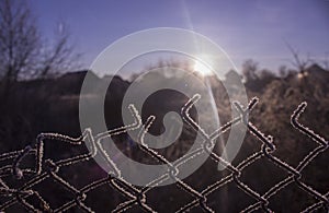 Torn fence of metal mesh covered with frost. The sun shines early in the morning on the frozen grid.