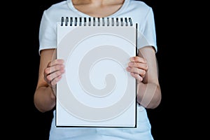 Torso of caucasian woman photographed against black background, holding up blank spiral bound note pad