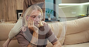 A tortured blond man with a beard and glasses sits on the sofa while his little albino son pesters him and wants to play