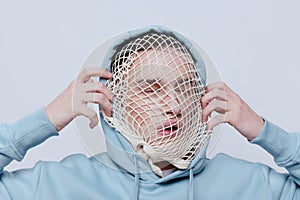 tortured, suffering from problems, a man with a mesh on his face stands in a light blue hoodie on a light background and