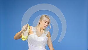 Tortured young beautiful blonde woman in a wedding white dress and veil on a blue background, takes off her shoes. The