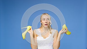 Tortured young beautiful blonde woman in a wedding white dress and veil on a blue background, takes off her shoes. The