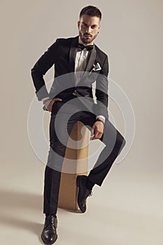 Tough groom holding hand in pocket, wearing tuxedo while sitting