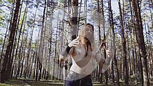 A tough guy in the woods throws a knife from hand to hand. Does the knife trick. The man has long hair and a braid on his beard.