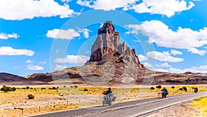 Touring Bike Riders passing the rugged peaks of El Capitan and Agathla Peak towering over the desert landscape of Monument Valley