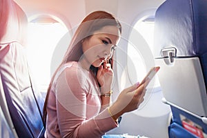 Tourist asian woman sitting near airplane window at sunset and using mobile phone during flight