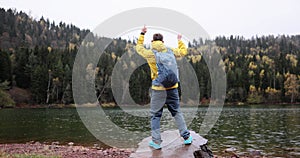 Tourist with backpack dances standing on rock against lake