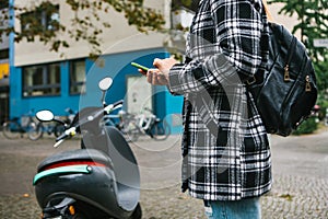 A tourist with a backpack is going to use an electric scooter through a mobile application in the phone and distally