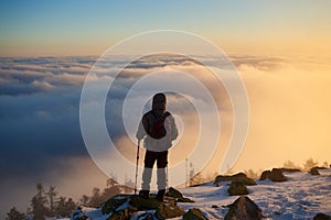 Tourist with backpack hiking on rocky mountain peak on background of foggy valley and blue sky at sunrise.