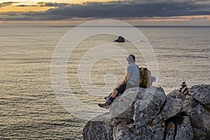 Tourist with a backpack sits on a rock looking at the sunset over the ocean