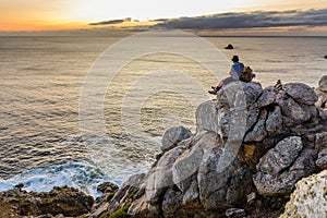 Tourist with a backpack sits on a rock looking at the sunset over the ocean