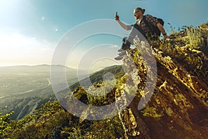 Tourist with backpack sits on top of the mountain and takes pictures of the landscape on cellphone