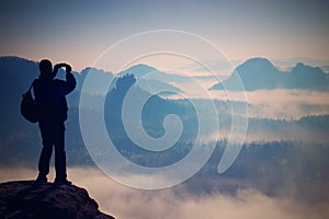 Tourist with backpack takes photos with smart phone on peak of rock. Dreamy fogy valley below