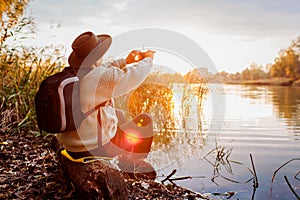 Tourist with backpack taking photos using smartphone of river at sunset. Woman travels admiring autumn nature