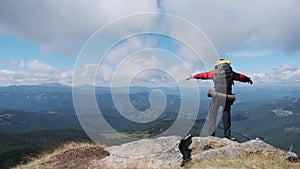 Tourist with Backpack on the Top of Mountain near the Cliff Raises Hands to Side