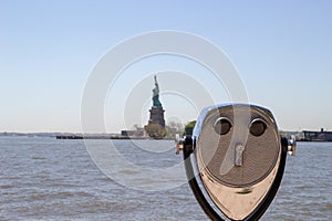 Tourist binoculars on Ellis Island with Statue of Liberty visible in the blurred background. There is a blue sky and water visible