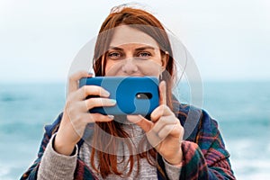 Tourist caucasian red hair woman takes pictures on smartphone on the beach near the ocean, travel vacation concept