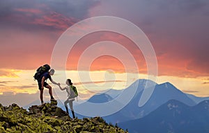 Tourist couple helping each other to climb high rock in evening mountains at sunset