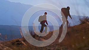 tourist couple man and woman with backpack walking and climbing up on top of mountain at sunset