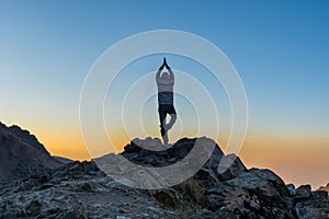 A tourist doing yoga on the climax of rocky mountain watching sun rising in Darband valley in autumn in dawn against colorful sky