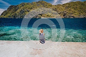 Tourist female rear view sitting pier edge on Matinloc island on hoping tour C. El Nido, Palawan, Philippines