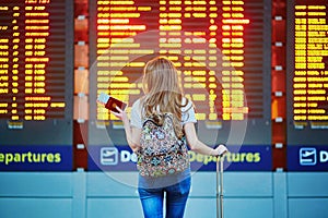 Tourist girl with backpack and carry on luggage in international airport, near flight information board