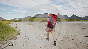 A tourist girl with a big red backpack and a hat walks along the seashore in the mountains. beautiful norway summer