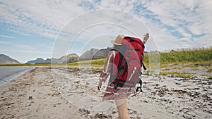 A tourist girl with a big red backpack and a hat walks along the seashore in the mountains. beautiful norway summer