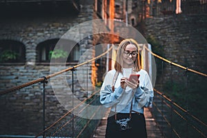 Tourist girl blonde with glasses uses smartphone  traveling through old historical city  Europe, young hipster plans walk