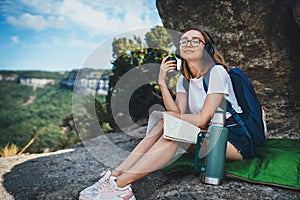 Tourist girl with closed eyes enjoying coffee from thermos and summer trip traveling in mountains, female hipster with glasses