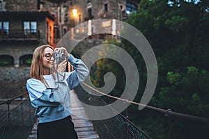 Tourist girl with glasses stands on suspension bridge on background historical city and takes photo with retro camera