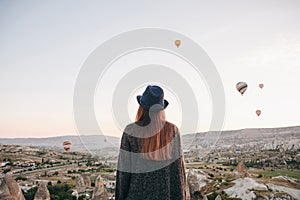 A tourist girl in a hat admires hot air balloons flying in the sky over Cappadocia in Turkey. Impressive sight.