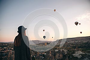 A tourist girl in a hat admires hot air balloons flying in the sky over Cappadocia in Turkey. Impressive sight.