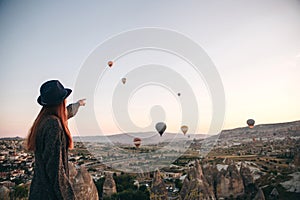 A tourist girl in a hat admires hot air balloons flying in the sky over Cappadocia in Turkey. Impressive sight.