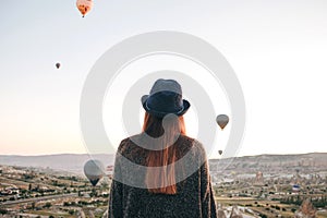 A tourist girl in a hat admires hot air balloons flying in the sky over Cappadocia in Turkey. Impressive sight.