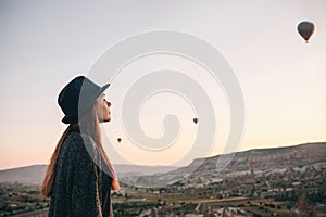 A tourist girl in a hat admires hot air balloons flying in the sky over Cappadocia in Turkey. Impressive sight.