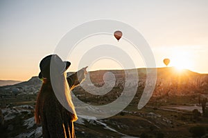 A tourist girl in a hat admires hot air balloons flying in the sky over Cappadocia in Turkey. Impressive sight.