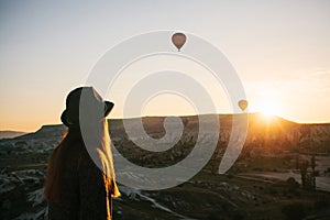 A tourist girl in a hat admires hot air balloons flying in the sky over Cappadocia in Turkey. Impressive sight.