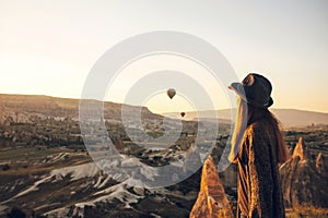 A tourist girl in a hat admires hot air balloons flying in the sky over Cappadocia in Turkey. Impressive sight.