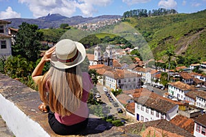 Tourist girl with hat sitting on wall looking at panoramic view of the historic city of Ouro Preto, Minas Gerais, Brazil