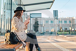 Tourist girl sits at public transport stop and looks in smartphone. Girl waiting for the bus.