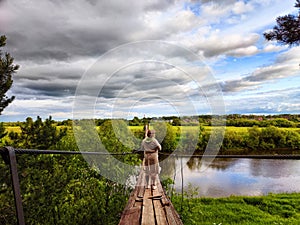 A tourist girl walks along an old wooden suspension bridge over the river and the sky with disturbing clouds. Suicide in