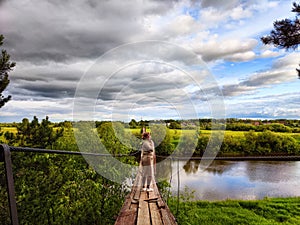 A tourist girl walks along an old wooden suspension bridge over the river and the sky with disturbing clouds. Suicide in